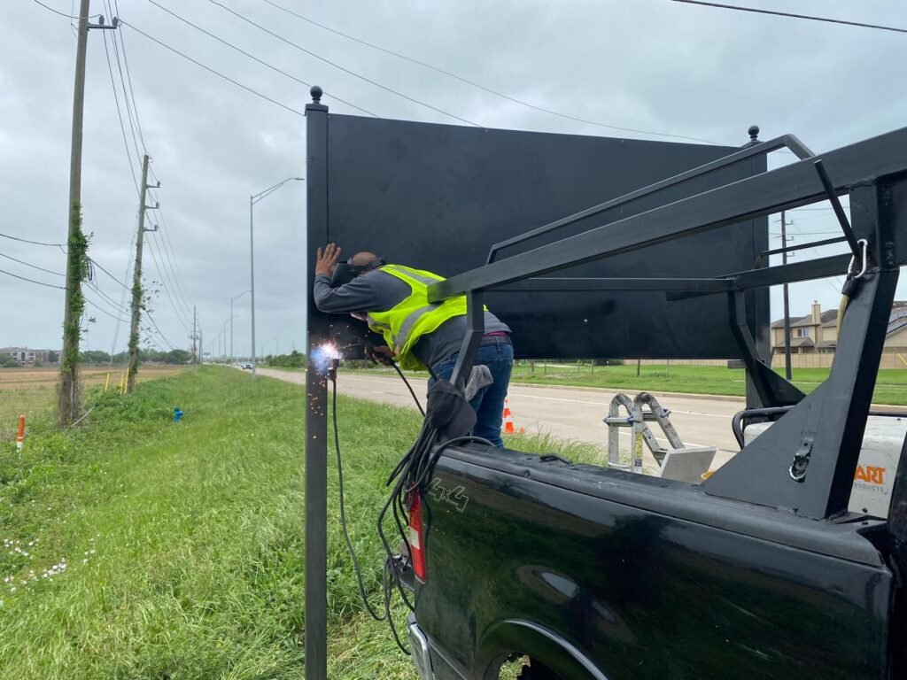 A professional welder repairing a large black metal privacy fence panel from the back of a work truck along a roadside.