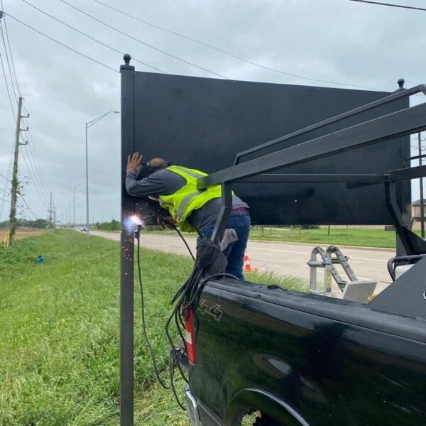 A professional welder repairing a large black metal privacy fence panel from the back of a work truck along a roadside.
