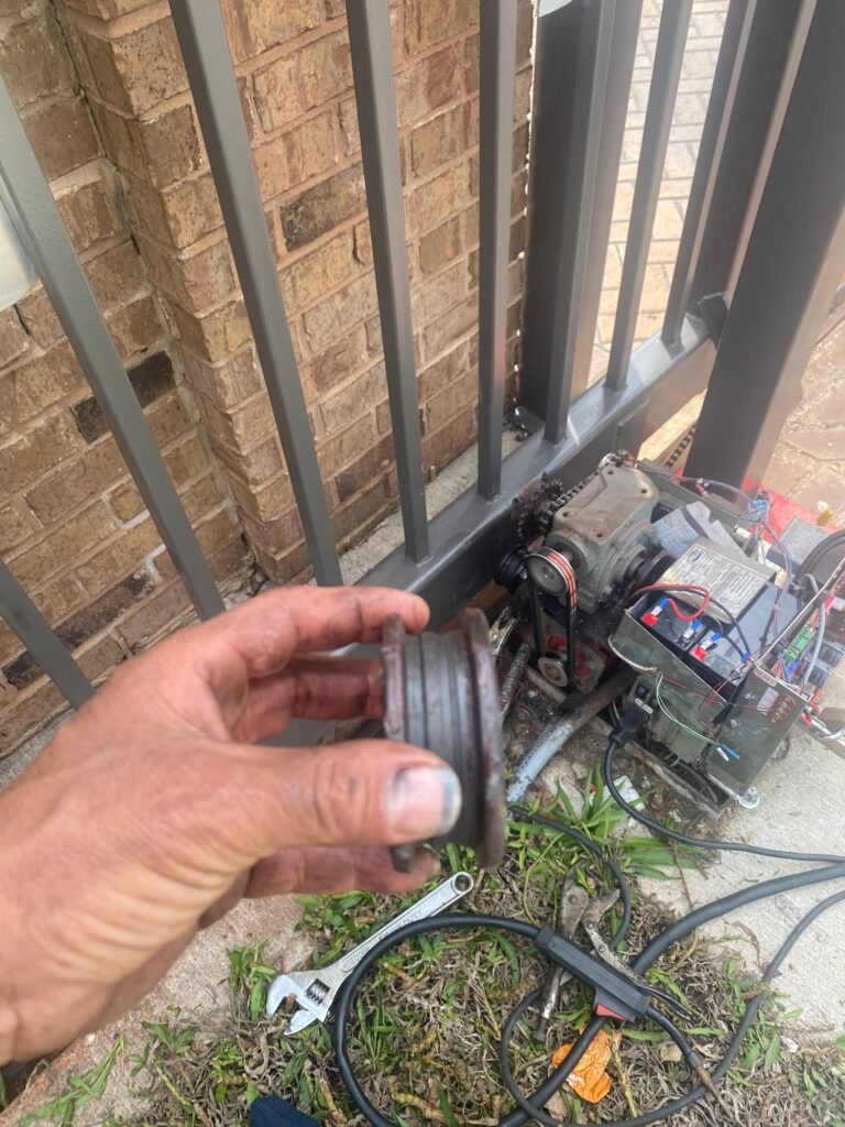 A technician holding a worn metal gate wheel during an electric sliding driveway gate track and motor repair.